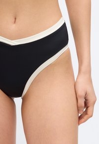 Γυναίκα με μαύρο μαγιό κάτω μέρος με λευκή επένδυση, εμφανίζοντας το κάτω μέρος της κοιλιάς, το πάνω μέρος των μηρών και το δεξί χέρι μπροστά σε απλό φόντο.