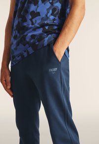 Camiseta de camuflaje azul con cuello redondo y mangas cortas, combinada con pantalones joggers azul marino que presentan un logo y bolsillos laterales.