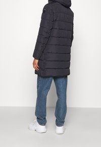 En puffig svart jacka med huva, med horisontell sömnad och en logotyp på axeln. Kombinerad med ljusblå jeans och vita sneakers.