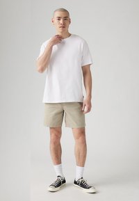 Cămașă albă din bumbac, pantaloni scurți bej deschis, șosete albe, adidași negri. Modelul stă cu o mână pe gât, păr scurt și fundal neutru.