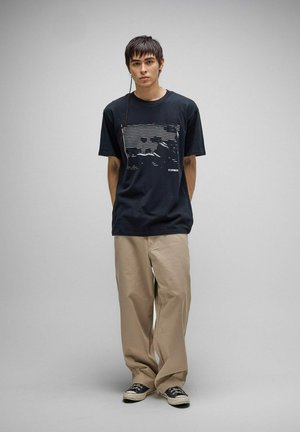 Jeune homme debout, les mains dans le dos, portant un t-shirt graphique noir, un pantalon beige à jambes larges et des baskets blanches et noires, fond neutre.