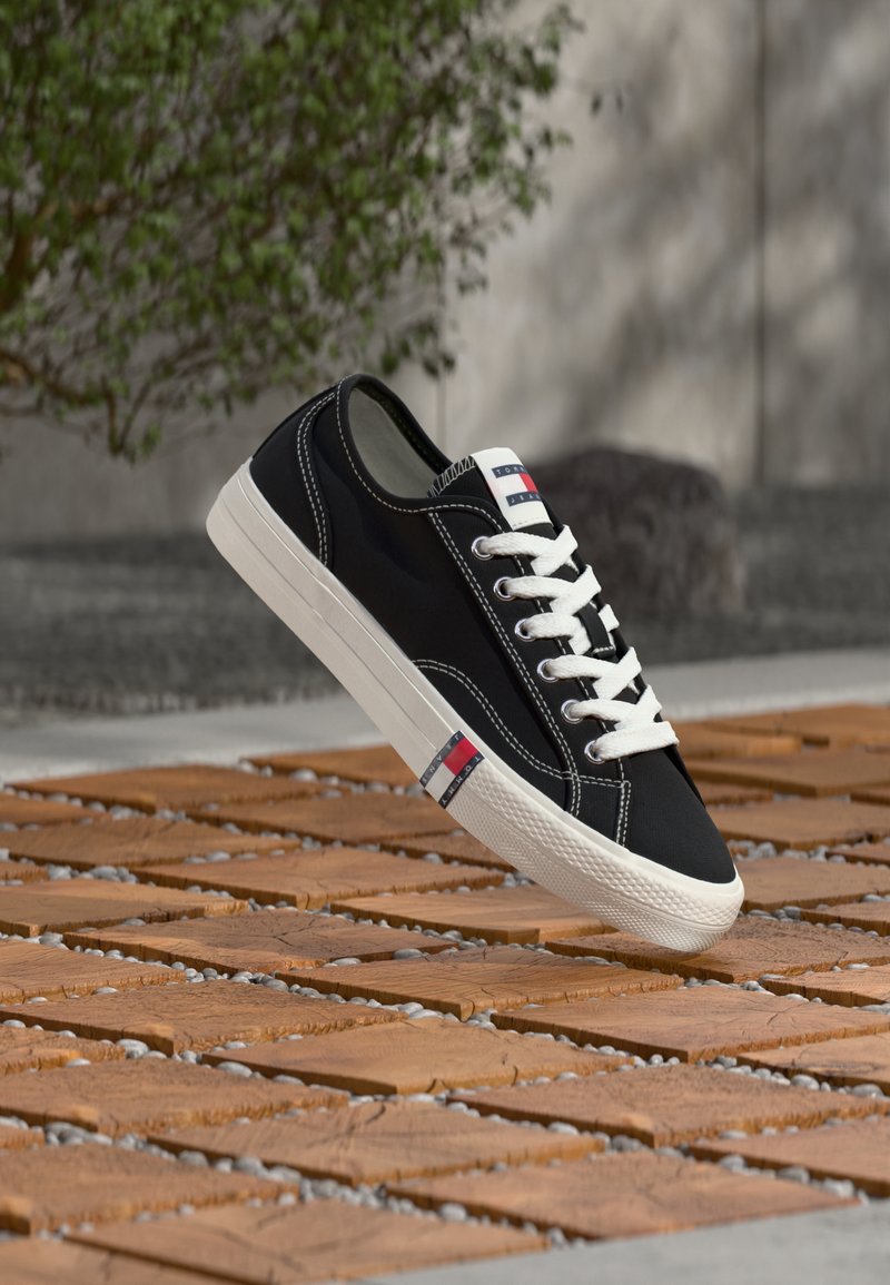 Pantof sport din pânză neagră cu șireturi și cusături albe, având un detaliu cu logo în dungi roșii, albe și albastre pe lateral, talpă din cauciuc.