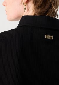 Zwarte geribbelde jas met een hoge kraag, voorzien van een gouden "Barbour International" label op de achterkant en textiel met structuur. Gouden oorbel zichtbaar.