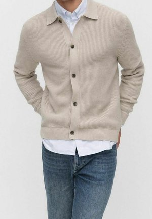 Cardigan en mailles beige boutonné porté sur une chemise à rayures bleu clair, associé à un jean bleu de lavage moyen.