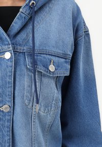 Τζάκετ τζιν σε ανοιχτό μπλε με κουκούλα, διαθέτοντας ασημί αξεσουάρ, μπροστινή τσέπη και σκούρες μπλε κορδόνια. Υφή υφάσματος με ορατές ραφές.