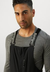Salopete negre cu bretele reglabile și un logo, închidere față cu fermoar și un tricou gri cu mânecă lungă, ajustat, dedesubt.