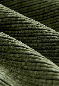 Grüner Cord-Stoff mit ausgeprägten Rillen, der eine weiche Textur und ein geripptes Muster zeigt. Das Material ist plüschig und samtig.