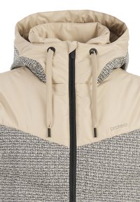 Bicolor-Jacke mit einem beigen oberen Bereich mit hohem Kragen und einem strukturierten, schwarz-weißen gemusterten unteren Bereich. Schwarzer Reißverschluss und Kordelzüge.