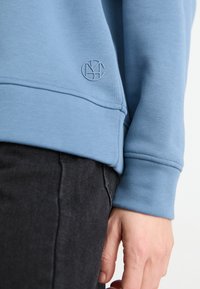 Modré mikina se žebrovanými manžetami a spodním lemem, s decentně vyraženým logem na přední spodní části vlevo. Vystavená s tmavými džíny.