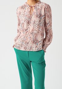 Blouse florale en rose, marron et bleu, avec un décolleté en trou de clé et des poignets froncés, assortie à un pantalon taillé vert. Tissu texturé.