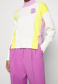 Pull-over en blanc, jaune, rose, avec un col montant côtelé et un logo "B" ; assorti à un pantalon rose vif. Texture lisse, design moderne.