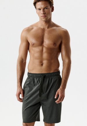 Młody mężczyzna o atletycznej sylwetce, ubrany w ciemnoszare spodenki ze sznurkiem, stoi na tle gładkiej białej ściany.