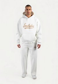 Vitt set med hoodie och mjukisbyxor, med en beige logotyp på framsidan. Mjukt material, avslappnad passform, med en känguruficka och ribbade muddar.