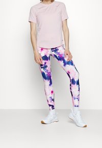 Top de manga corta rosa claro combinado con leggins multicolores que presentan un patrón de acuarela en rosa, púrpura y azul, completando el look atlético.