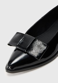 Pantofi negri cu vârf ascuțit, cu un finisaj patent neted și un accent de fundiță texturată pe față. Interiorul este negru și elegant.