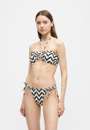 Młoda kobieta ubrana w dwuczęściowy bikini w czarno-białe wzory z halterem i bokami z wiązaniami, stojąca na jednolitym tle.