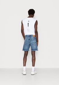 Mies, który nosi białą koszulkę bez rękawów z czarnym nadrukiem, postrzępione niebieskie spodenki dżinsowe, białe skarpetki i białe trampki, stojący na białym tle.