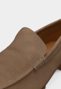 Καφέ σουέντ παπούτσι slip-on με ορατή ραφή και στρογγυλεμένη πάνω άκρη σε ουδέτερο φόντο.