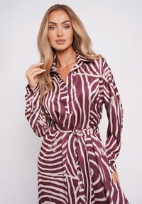 Kobieta z długimi blond włosami w maroon i białą sukience w paski zebry z paskiem i kołnierzykiem, pozująca na jednolitym jasnym tle.