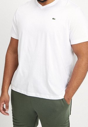White cotton t-shirt with a v-neck, short sleeves, and a small green Lacoste logo on the chest. Textured fabric, relaxed fit.