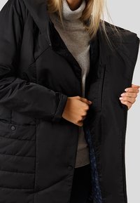 Osoba v černém prošívaném kabátu přes světle šedý rolák, která drží kabát otevřený, aby ukázala jeho vnitřní podšívku.