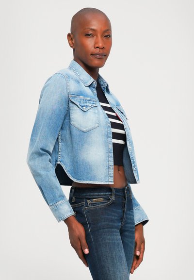 Woman with shaved head wearing a light blue cropped denim jacket over a black and white striped crop top and dark blue jeans against a plain background.