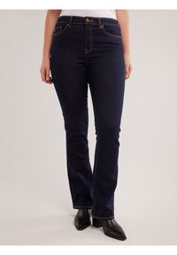 Žena u tamnoplavim trapericama zvonolikog kroja s narančastim šavovima i crnim čizmama s potpeticom, stoji uza svijetlu pozadinu.