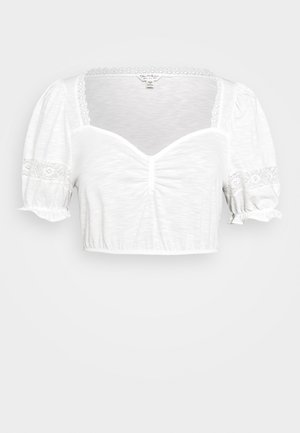 Blouse blanche courte avec manches bouffantes, bordure en dentelle sur l'encolure et les manches, et détail froncé au centre devant.