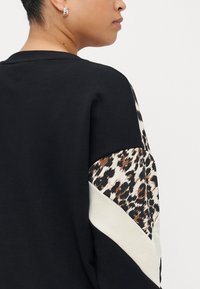 Juodas megztinis su leopardų raštu akcentais rankovėse, su kontrastinga kreminės spalvos įstriža dalimi. Minkšta medžiaga, laisva forma.