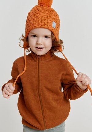Orange Strickmütze mit Bommel; braune Jacke mit Reißverschluss und hohem Kragen; Kind hält die Mützenbändel; hellblaue Jeans; neutraler Hintergrund.