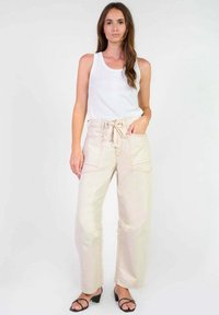 Pantalons beige taille haute avec une coupe ample et une ceinture à nouer. Dotés de deux poches avant et de détails de couture visibles. Portés avec un débardeur blanc.