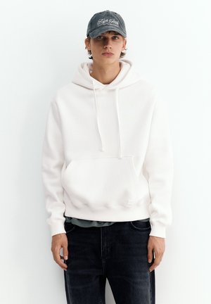 Un jeune homme pose avec un sweat à capuche blanc uni, un jean foncé et une casquette grise sur un fond de studio clair.