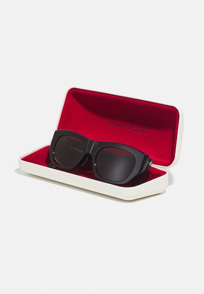 Occhiali da sole rettangolari neri con lenti scure, posizionati in un astuccio bianco con un interno rosso. L'interno dell'astuccio mostra il nome del marchio, Alexander McQueen.