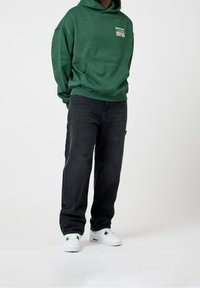 Grüner Kapuzenpullover mit Vordertasche, kombiniert mit dunklen Denim-Jeans und weißen Sneakern mit grünen Akzenten. Einfaches und lässiges Outfit.