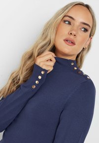 Marinefarbener Rollkragenpullover mit langen Ärmeln, ausgestattet mit goldenen Knopfdetails an einer Schulter, gefertigt aus einem strukturierten Strickstoff.