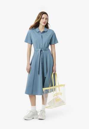 Blue polo dress with short sleeves, buttons, and a waist tie. Clear tote bag with yellow accents and logo. White sneakers complete the look.