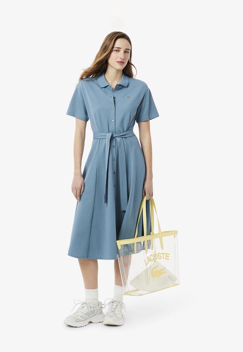 Abito polo blu con maniche corte, bottoni e cintura in vita. Borsa tote trasparente con accenti gialli e logo. Sneakers bianche completano il look.