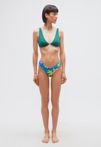 Γυναίκα με κοντά σκούρα μαλλιά, φορώντας πράσινο τοπ μπικίνι και ζωντανά φλοράλ κάτω μέρος μπικίνι, όρθια ξυπόλητη σε απλό λευκό φόντο.