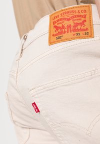 Jeans in denim beige con una toppa in pelle sulla vita con branding rosso e arancione. Cuciture distintive sulle tasche posteriori e una piccola etichetta rossa.