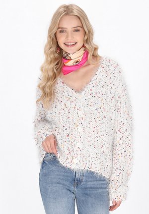 Cardigan blanc en peluche avec des touches multicolores, col en V et fermeture à boutons, associé à un jean bleu et à un foulard enroulé rose à motifs.