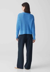 Maglione blu lavorato a maglia con un motivo testurizzato, leggermente accorciato sul retro, abbinato a pantaloni larghi scuri e calzature chiare.