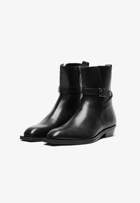 Botas de cuero negras hasta el tobillo con punta afilada, cremallera lateral y una correa decorativa con una hebilla de plata. Textura lisa y tacón bajo.