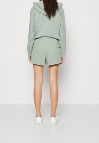 Sweat à capuche vert clair avec cordon de serrage et poche kangourou, assorti à un short. Le short est doté de poches latérales. Des baskets blanches complètent la tenue.