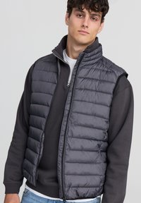 Szary kamizelka puchowa z wysokim kołnierzem i zamkiem błyskawicznym, z pikowanym wykończeniem i lekką konstrukcją. Noszona na ciemnej bluzie.