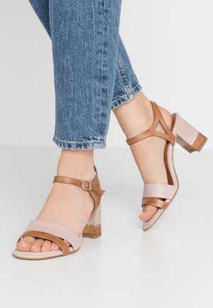 Des sandales à talons blocs beiges et marron présentent de larges brides avant, une sangle cheville avec une boucle, et une finition lisse avec un accent brillant au niveau du talon.