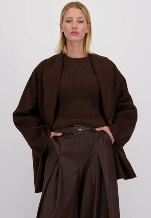 Vrouw met blond haar, gekleed in een donkerbruin gebreid sweater, brede riem, geplooide leren broek en een oversized bruine jas, met haar handen in de zakken.