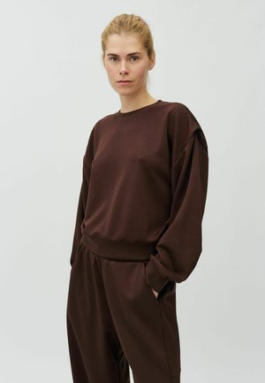 Kvinde med lyst hår sat tilbage iført løs brun sweatshirt og matchende bukser med hænderne i lommerne mod ensfarvet baggrund.
