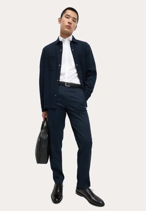 Marynarska kurtka na białej koszuli, w połączeniu z ciemnoniebieskimi spodniami i czarnymi skórzanymi butami, trzymająca czarną skórzaną torbę.