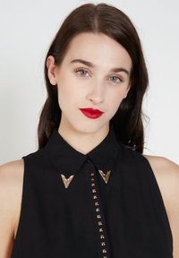 Joven mujer con cabello largo y oscuro, lápiz labial rojo, vestida con una camiseta sin mangas negra con puntas de cuello en forma de V de metal y adornos de tachuelas.