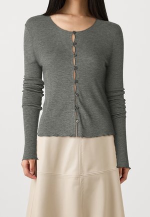 Nainen pukeutuneena istuvaan harmaaseen ribbineuleiseen neuletakkiin, jossa epätasainen nappisuljin, ja beige nahkahame.